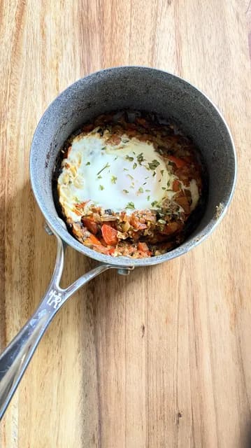 A poached egg nestled in a spiced tomato and pepper stew, garnished with fresh herbs in a skillet.