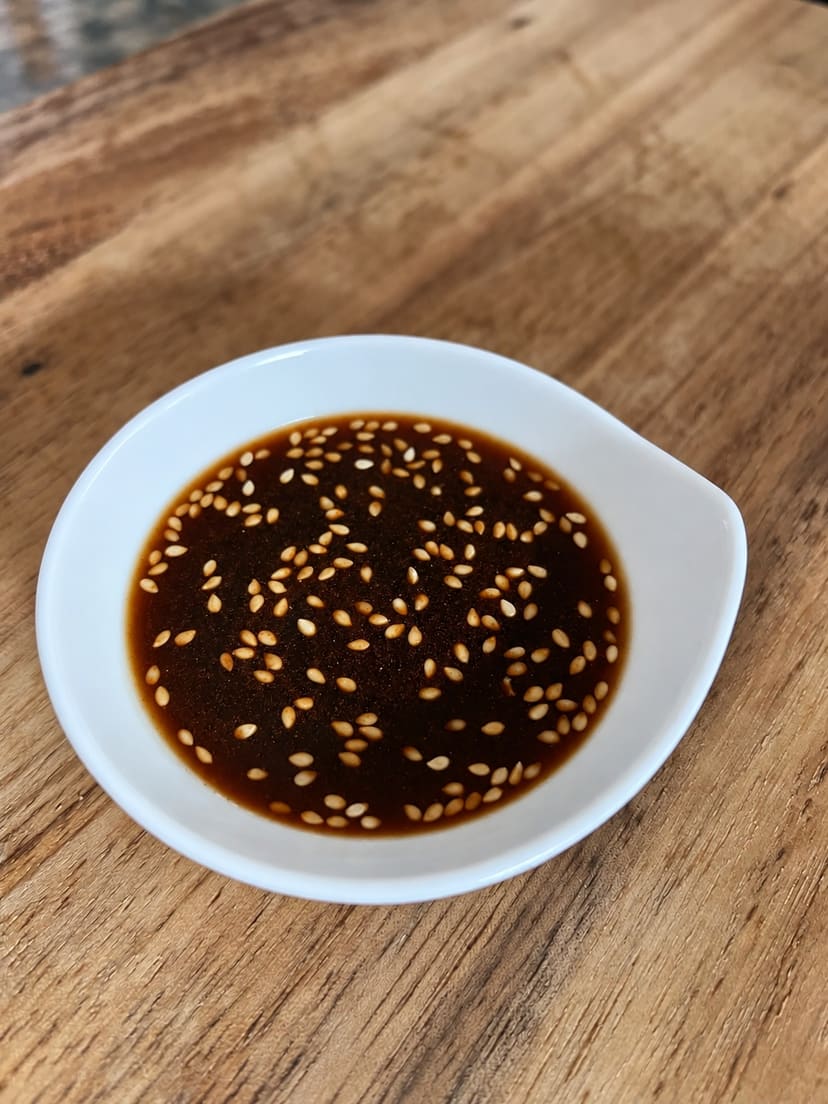 A small white bowl filled with a dark soy-based sauce garnished with visible sesame seeds and red pepper flakes.