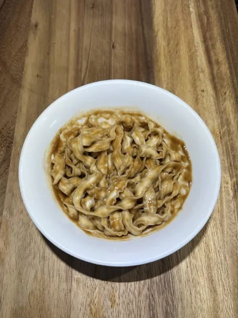 Bowl of wheat noodles coated in a rich peanut butter sauce, garnished with toasted sesame oil.